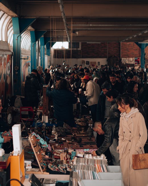 A crowded indoor market scene with numerous people browsing and purchasing items from various stalls. The space features high ceilings with exposed beams and large windows allowing natural light to filter in. Market tables are filled with a diverse array of goods, including books, textiles, and miscellaneous objects, some of which are stacked or laid out flat. The colours within the scene range from earthy tones of the brick wall to the vibrant decorations hanging from the ceiling. Several individuals appear engaged in examining items closely, while others stand in small groups or move through the space. The environment is lively but orderly, typical of a market where people are handling various types of goods, some possibly for resale or personal use. The scene illustrates a typical setting where comprehensive waste management and rubbish removal, such as through private collection services like Waste Clearance Tooting, might be relevant for clearing out unsold stock or old items afterward, reflecting the practical aspect of managing waste from such environments.