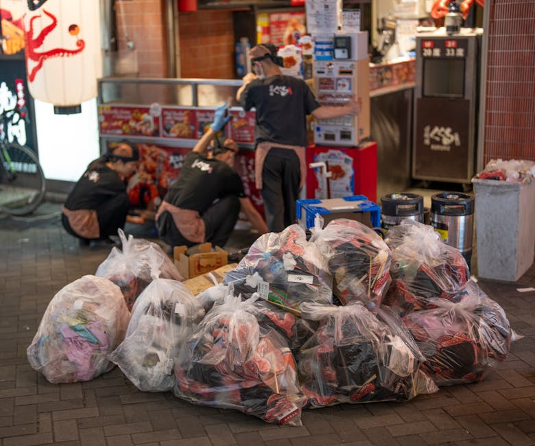 A large pile of transparent plastic rubbish bags filled with mixed waste, including textiles and packaging, is situated on a paved sidewalk in front of a commercial food outlet at night. Behind the bags, three workers dressed in black uniforms with brown aprons are actively engaged in clearing or organizing waste, with one standing and leaning over a counter, likely handling more waste or supplies. The scene is illuminated by ambient street lighting, and the background features a red-brick building with illuminated signage and a nearby trash bin. Visible equipment includes a blue plastic container, two black insulated bins, and packaged food displays, indicating an environment of on-site waste collection related to a food or retail business. This setting exemplifies private rubbish management by Waste Clearance Tooting, highlighting alternative waste handling outside of council services, with an emphasis on prompt, organized rubbish removal for commercial premises.