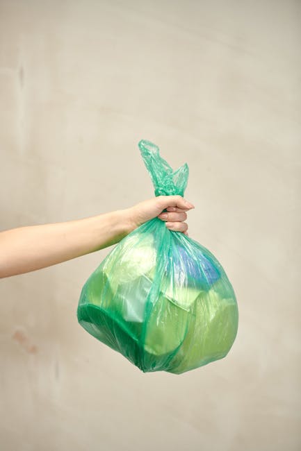 A human hand extends from the left side of the image, gripping a green plastic bag filled with waste. The bag appears to contain light-colored and possibly recyclable materials, with some parts of it showing hints of other colours like blue or darker shades. The bag is tied at the top with a twist, creating a gathered knot, and is held against a plain, neutral-toned background with subtle shading that suggests an indoor environment. The bag’s plastic surface is slightly translucent and glossy, reflecting ambient light. The overall scene emphasizes the act of waste collection or disposal, typical of rubbish removal services, and subtly aligns with private waste handling. The focus is on the bag, with the hand and arm occupying the left portion of the frame, and the bag’s rounded shape and gathered top are prominent. This visual can relate to independent collection or waste management activities handled by companies such as Waste Clearance Tooting, with an emphasis on rubbish and waste disposal processes.