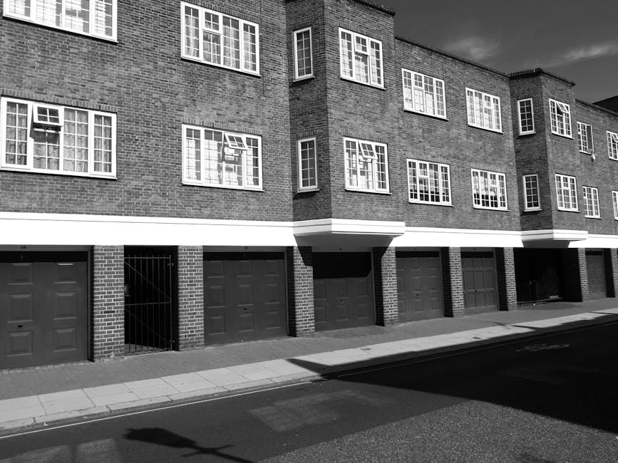 A black-and-white photograph depicting the ground level of a modern, multi-storey residential building with brick exterior walls. The building features several evenly spaced, rectangular windows with white frames, some partially open, revealing interior reflections. The lower portion of the building shows a row of closed garage doors, each painted in a dark tone, with some doors having slight variations in design. A narrow metal gate is visible between two of the garage doors, leading into a small enclosed area. The structure casts a shadow onto the sidewalk and street below, with a clear, sunny sky above and no vehicles or pedestrians present. The overall scene presents a clean, urban environment suited to private waste collection and rubbish removal services, with the building's facade and driveway prepared for managing refuse disposal related to residential property management. Waste Clearance Tooting may assist with on-site clearance or alternative waste handling for such properties, as suggested by the scene's context.