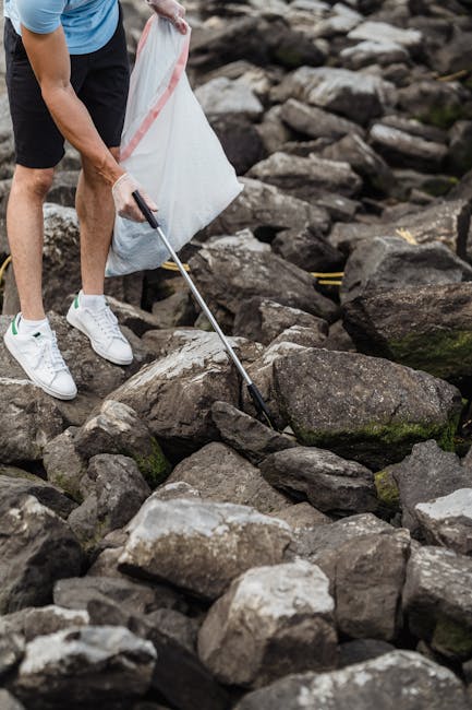 A person wearing white sneakers, black shorts, and a blue shirt is collecting rocks with a grabber tool while holding a large white trash bag on rocky terrain. The rocks vary in size and color, mostly shades of grey and brown with some patches of green moss. The scene appears to be outdoors in a natural setting, possibly near a shoreline or a riverbank, with the individual engaged in debris or waste removal. The person is positioned on a flat area among scattered rocks, with the background showing more rocks and natural landscape, consistent with a site where independent waste collection or environmental cleanup might be taking place, supported by services such as Waste Clearance Tooting that handle rubbish removal in various environments.