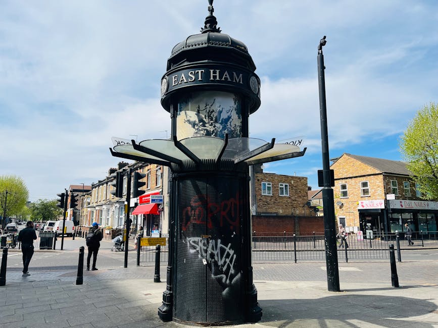 A freestanding cylindrical structure with a rounded dome top, marked with a sign reading 'EAST HAM', situated at a busy urban intersection. The structure features a black, metal mesh body with graffiti tags on its surface, and a decorative, slightly domed top with an ornate finial. Surrounding the structure are several black bollards, a tall streetlight to the right, and a row of shops and residential buildings in the background. Pedestrians are walking across the pavement, some wearing jackets, and the sky above is mostly blue with scattered clouds. The scene is well-lit with natural daylight, and the environment suggests a central town or neighborhood with good visibility of local amenities and public spaces, which aligns with the context of independent waste clearance or rubbish removal services in the area, as provided by Waste Clearance Tooting.