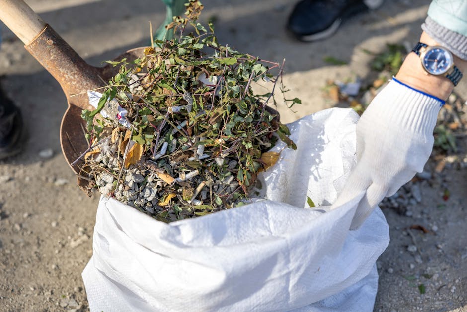 A person wearing a white glove and a wristwatch is lifting a white waste sack filled with garden debris, including small shrub branches, leaves, and gravel. The sack appears to be made of woven plastic material, securely holding the organic and inorganic plant material. In the background, part of a shovel with a rusted metal blade and wooden handle is visible, indicating activity related to clearing or loading waste. The scene is set outdoors on a gravel or dirt surface, with natural daylight illuminating the area. The presence of garden waste mixed with small stones in the sack relates to private or alternative waste disposal services, typical in rubbish removal or clearance operations by companies like Waste Clearance Tooting. The environment and tools suggest an on-site clearance process, supporting professional rubbish collection for garden or outdoor space cleanups.